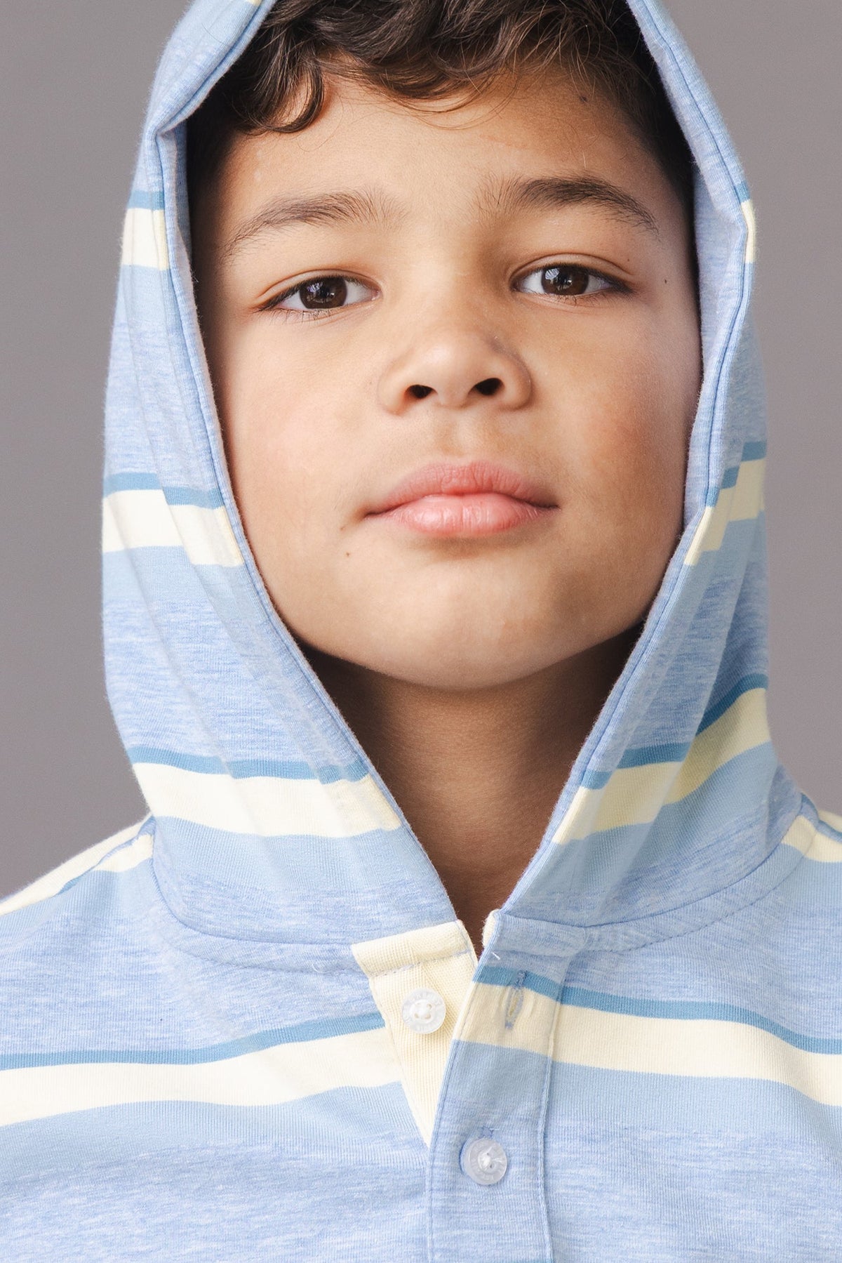 A young boy models a light blue hoodie with yellow and white stripes and a cozy hood. The hoodie features a Henley button placket, perfect for a stylish, casual look.