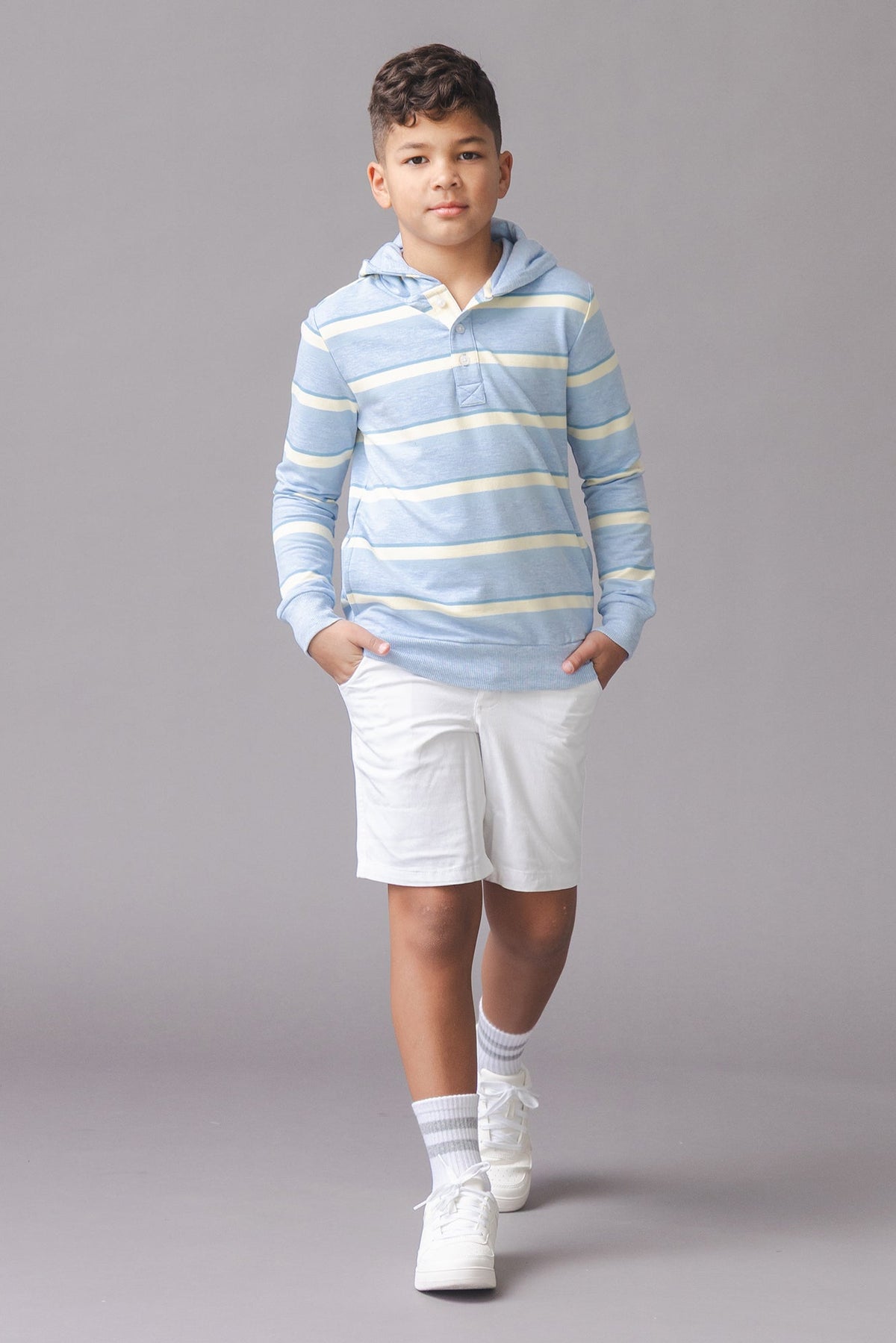 A child walks confidently in a light blue striped hoodie with a cozy hood and Henley button placket, paired with white shorts and sneakers, set against a neutral background.