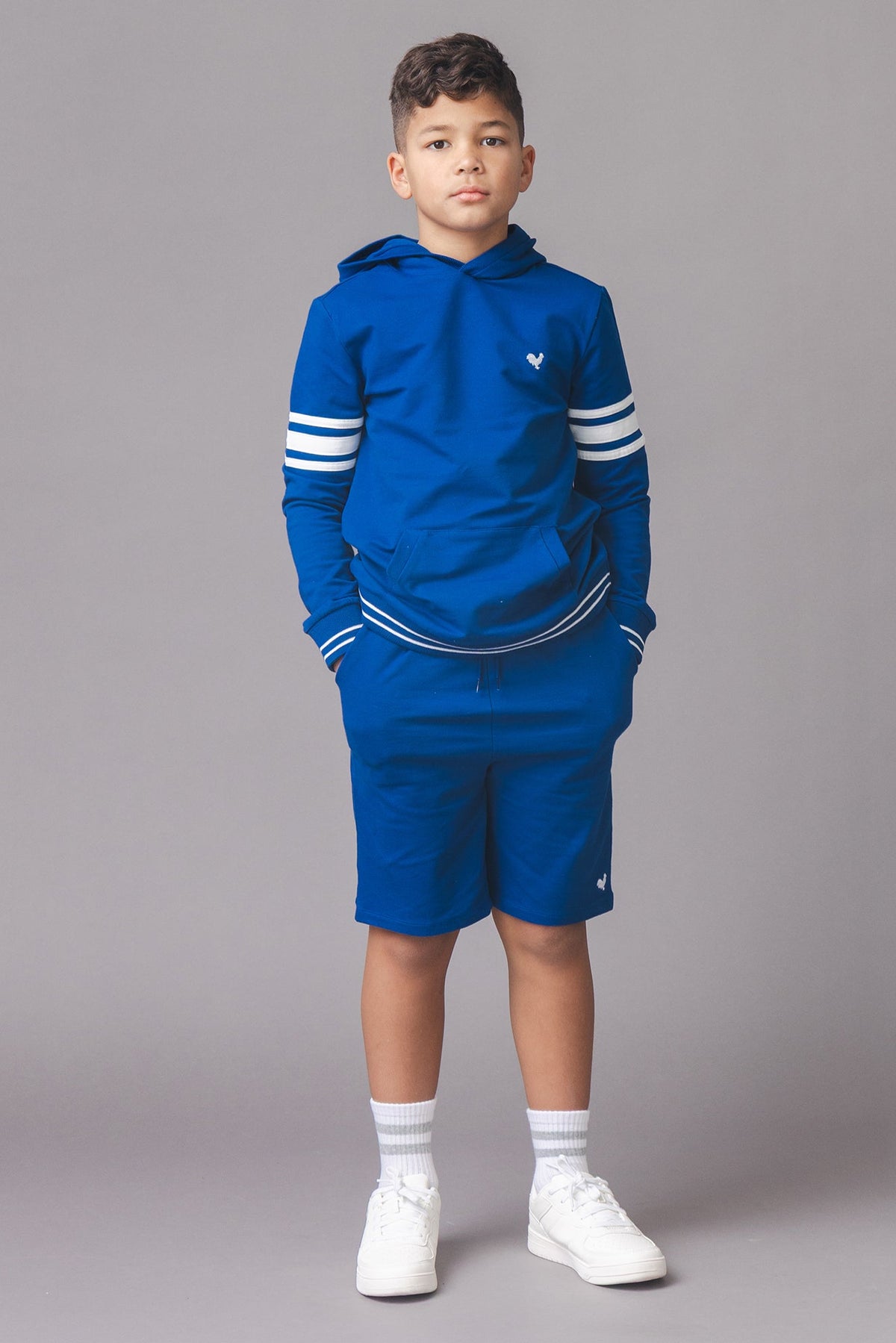 A boy stands against a gray backdrop, wearing a blue hoodie with white stripes and matching shorts. The outfit features a relaxed fit, perfect for casual play and comfort.