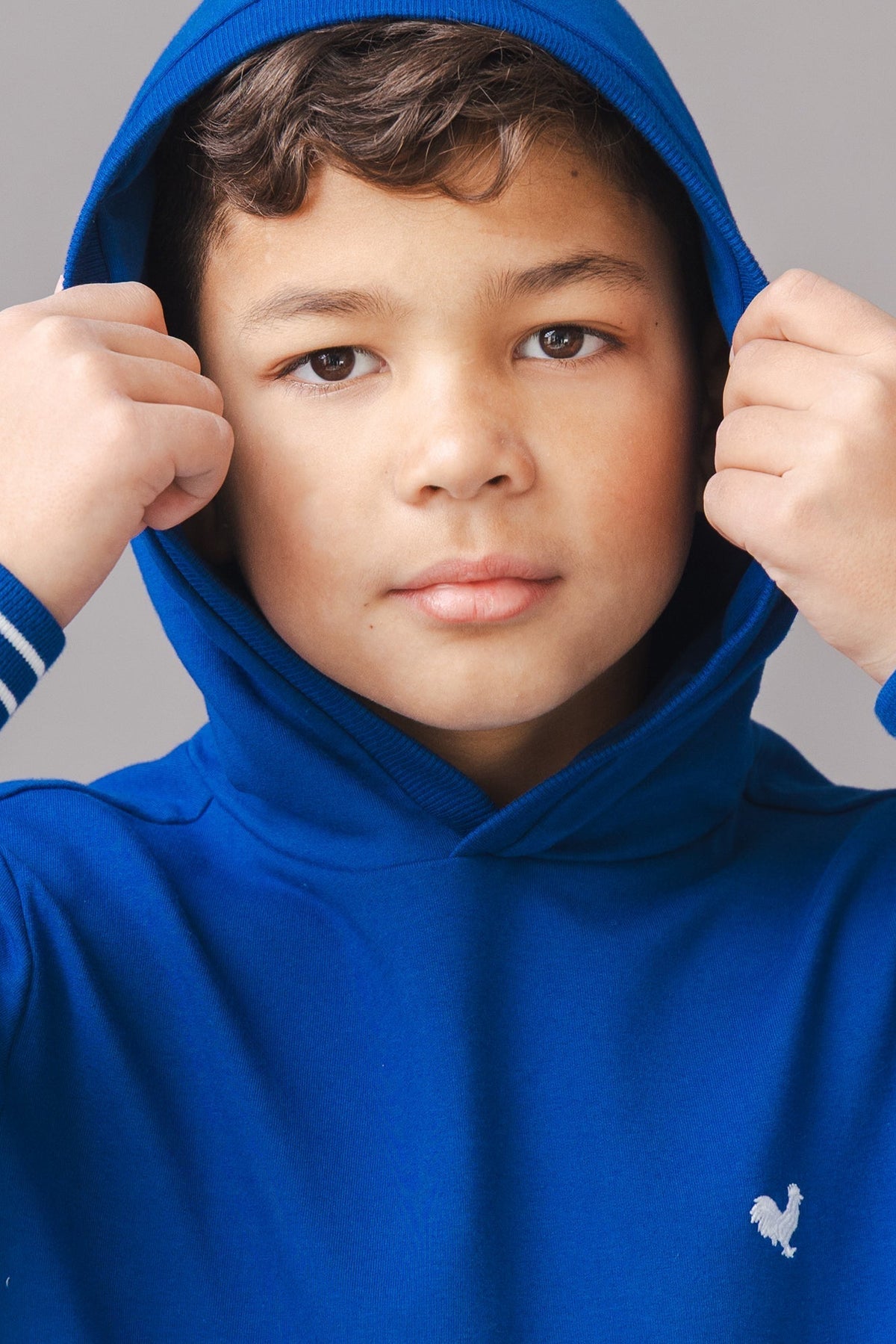 A young boy adjusts the hood of a vibrant blue hoodie, featuring white stripes on the sleeves and a small logo.