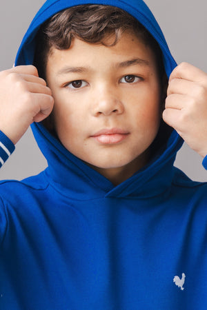 A young boy adjusts the hood of a vibrant blue hoodie, featuring white stripes on the sleeves and a small logo.