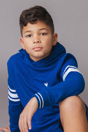 A young boy sits against a gray backdrop, wearing a vibrant blue organic cotton hoodie with white stripes on the sleeves.