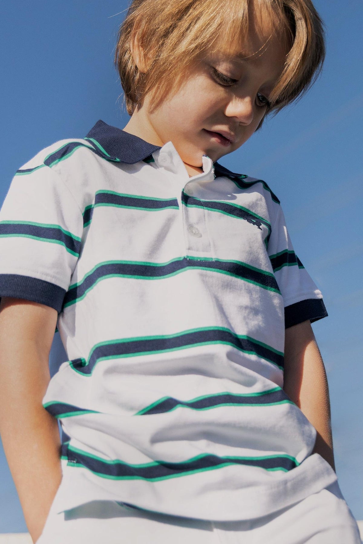 A young boy stands outdoors, wearing a white organic cotton polo shirt with green and navy stripes.