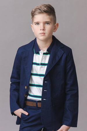 A young boy stands confidently wearing a navy blazer over a striped organic cotton polo. His hairstyle is neatly styled, and he poses against a neutral gray background.
