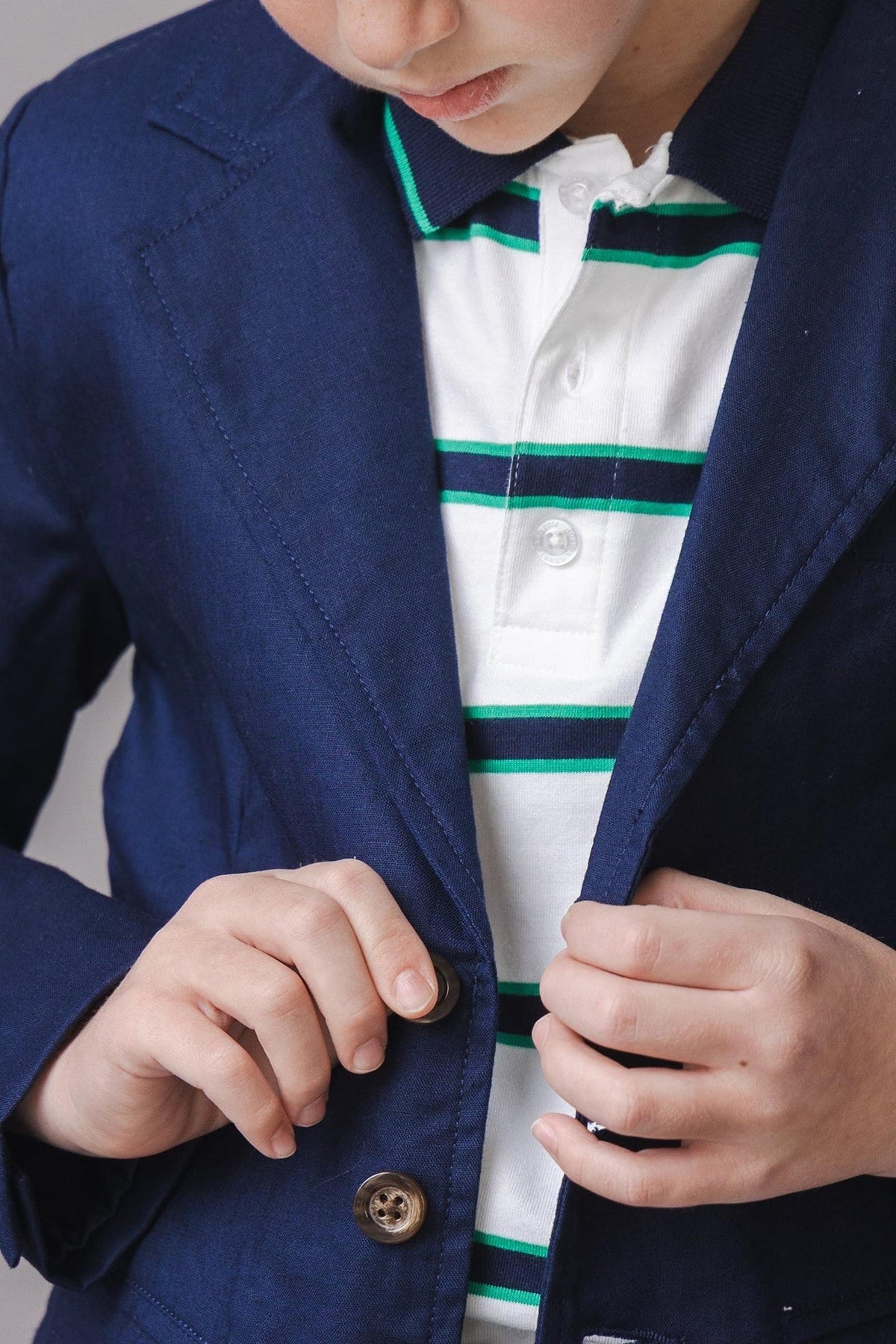 A young boy adjusts the buttons on a navy blazer over a white polo shirt with green and blue stripes. The outfit combines classic style with modern comfort.
