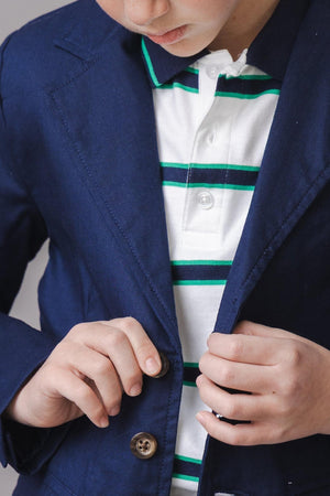A young boy adjusts the buttons on a navy blazer over a white polo shirt with green and blue stripes. The outfit combines classic style with modern comfort.