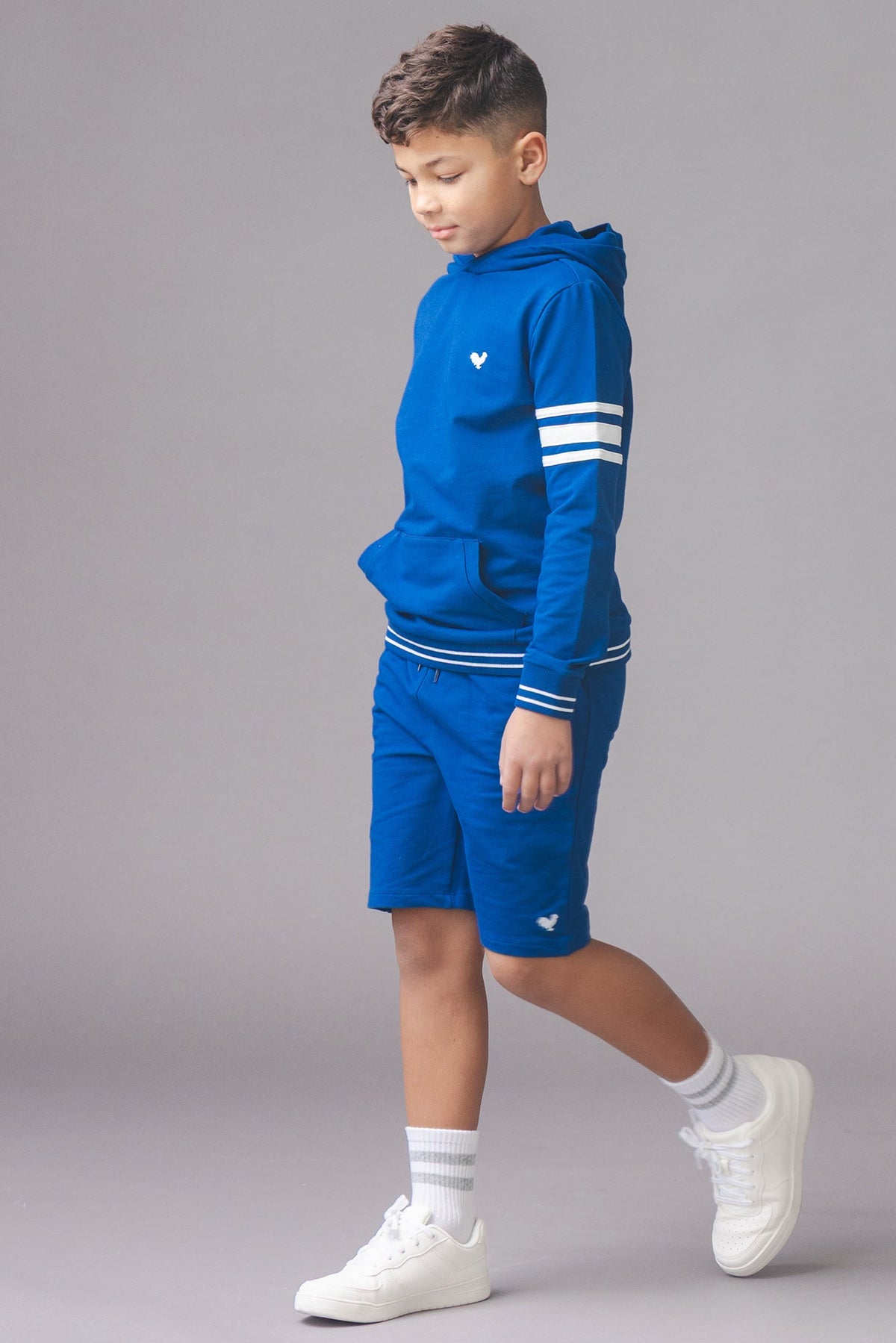 A young boy walks in a blue hoodie with white stripes and matching blue shorts. The outfit is made from soft fabric, ideal for active play and comfortable wear.