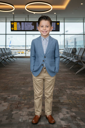 A young boy in a blue chambray blazer and beige pants stands smiling in an airport terminal.