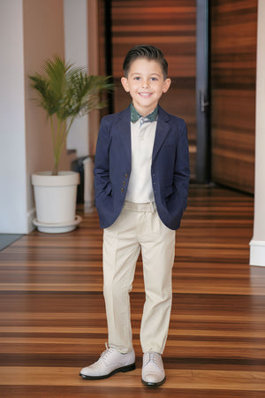 A young boy is smiling in a navy suit jacket, beige pants, and stylish shoes, standing in a modern indoor.