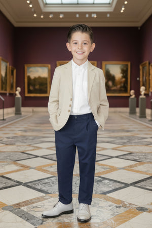 A young boy stands confidently in a museum, wearing a beige linen suit jacket over a white shirt and navy pants, complemented by stylish gray shoes.