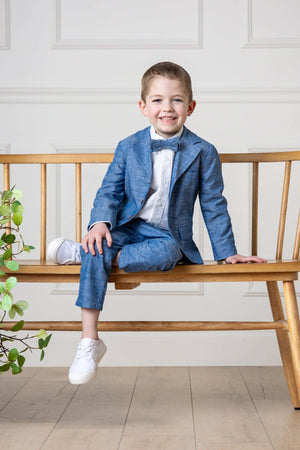 A young boy sits on a wooden bench wearing a blue suit, white shirt, and white sneakers, smiling happily.