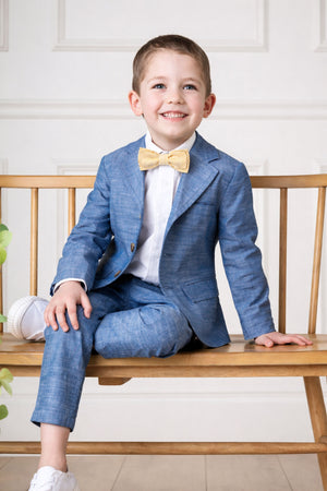 A young boy sits on a wooden bench, wearing a blue suit and a yellow striped bow tie, smiling brightly.