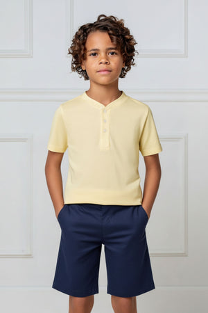A boy wears a pale yellow organic cotton short sleeve henley t-shirt, paired with navy shorts, against a neutral background.