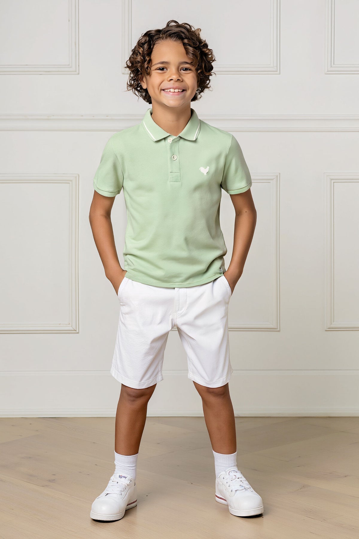 A young boy stands confidently wearing a soft green organic cotton polo shirt with a flat-knit collar, paired with white shorts and sneakers, set against a light-colored background.