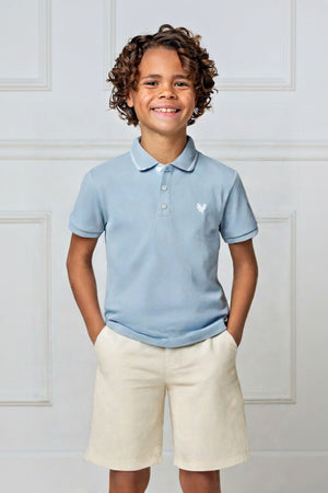 A smiling child wears a pale blue organic cotton polo shirt with a flat-knit collar, paired with light beige shorts.