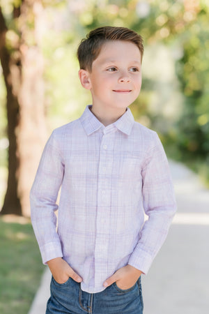 A smiling boy stands outdoors, wearing a light purple plaid button-down shirt and blue jeans, with hands in pockets.