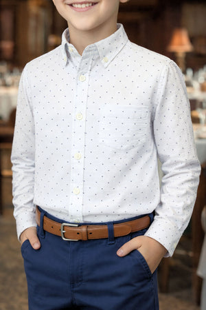A smiling boy wears a white seersucker shirt with blue dots and a brown belt, standing in a dining setting.