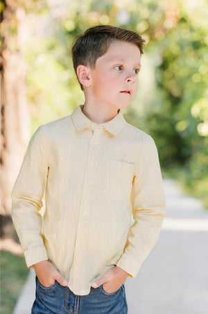 A young boy stands outdoors wearing a light yellow striped long sleeve shirt, with his hands in his pockets.