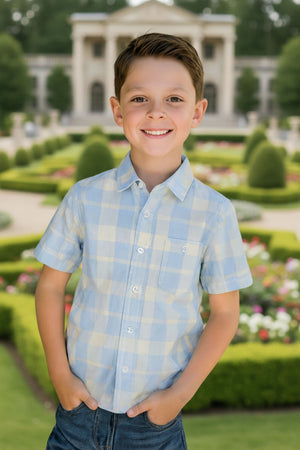 A boy in a light blue plaid short sleeve shirt smiles in a garden with a historic building behind.