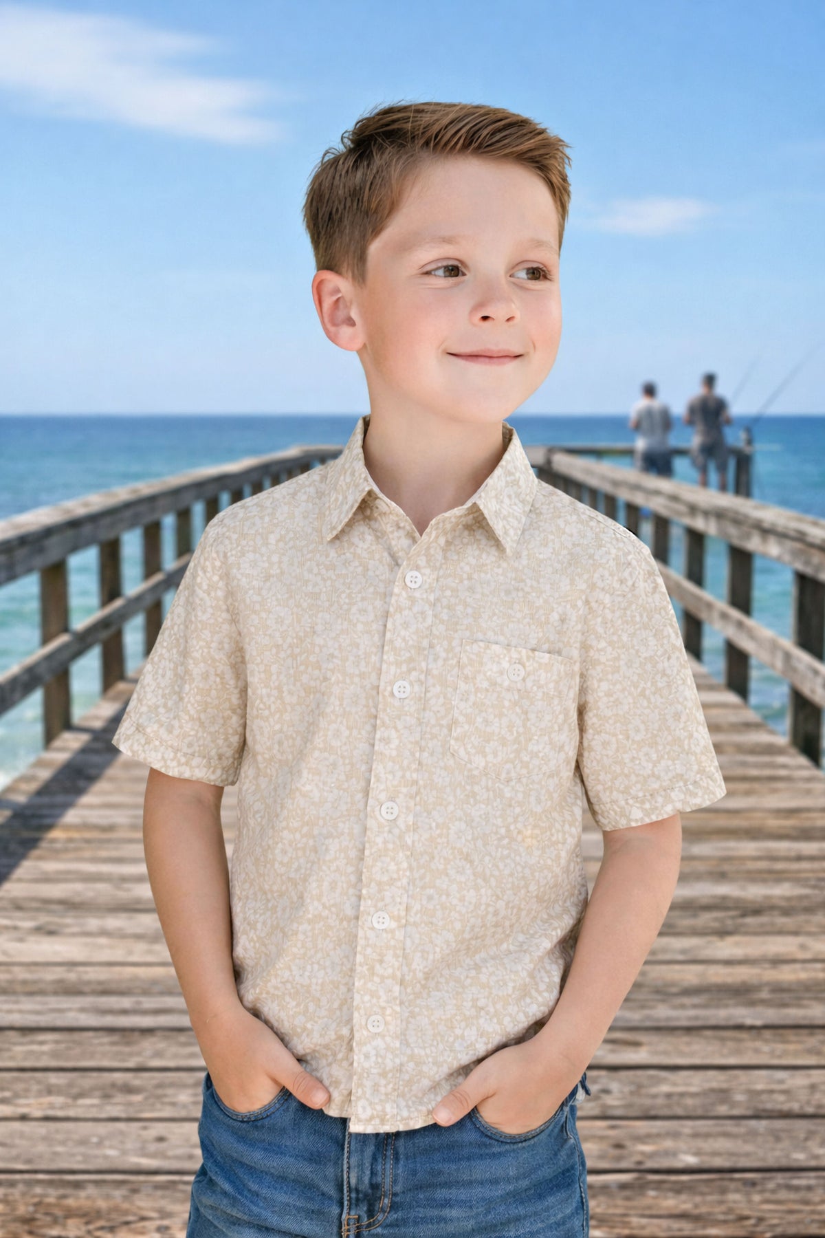 A young boy smiles while wearing a sandy floral short sleeve shirt, standing on a wooden pier by the ocean.