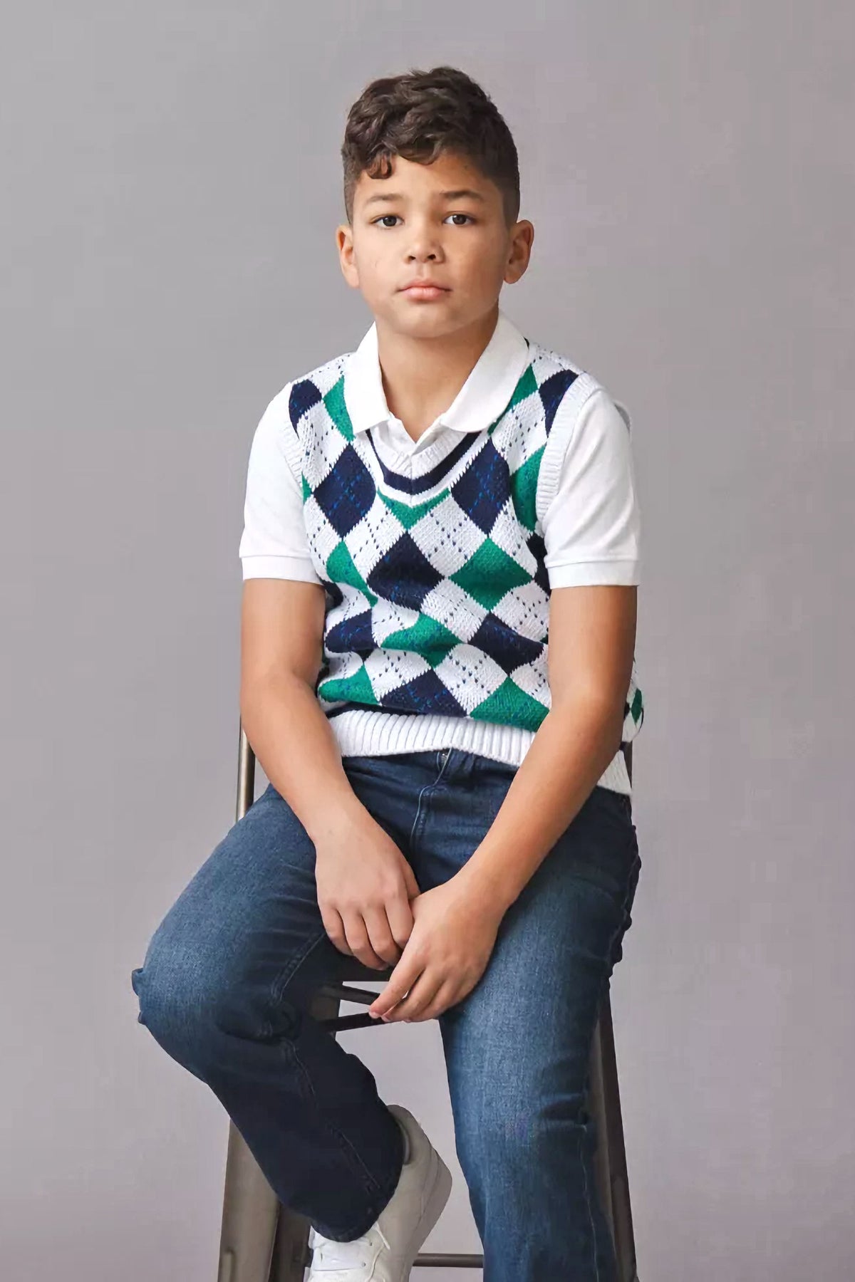 A young boy sits on a stool wearing a white collared shirt beneath a green and navy argyle sweater vest. He pairs it with dark jeans and white sneakers.
