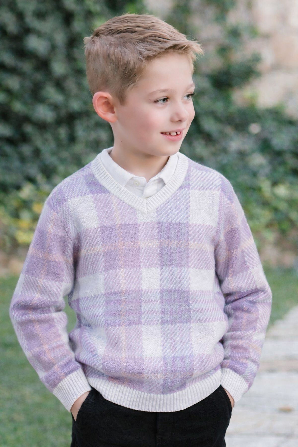 A young boy smiles while wearing a light purple plaid v-neck sweater made from organic cotton, paired with a white.