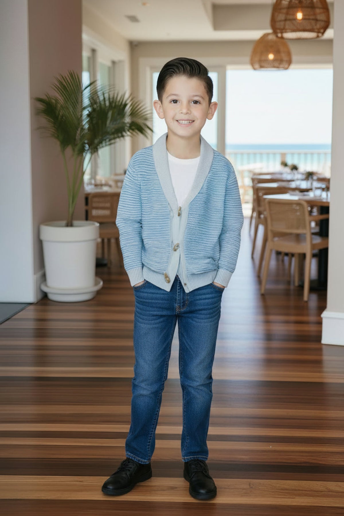 A smiling boy stands in a bright, airy room wearing a light blue cardigan, white shirt, and blue jeans.