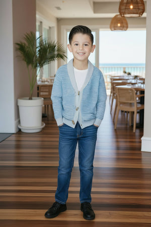 A smiling boy stands in a bright, airy room wearing a light blue cardigan, white shirt, and blue jeans.