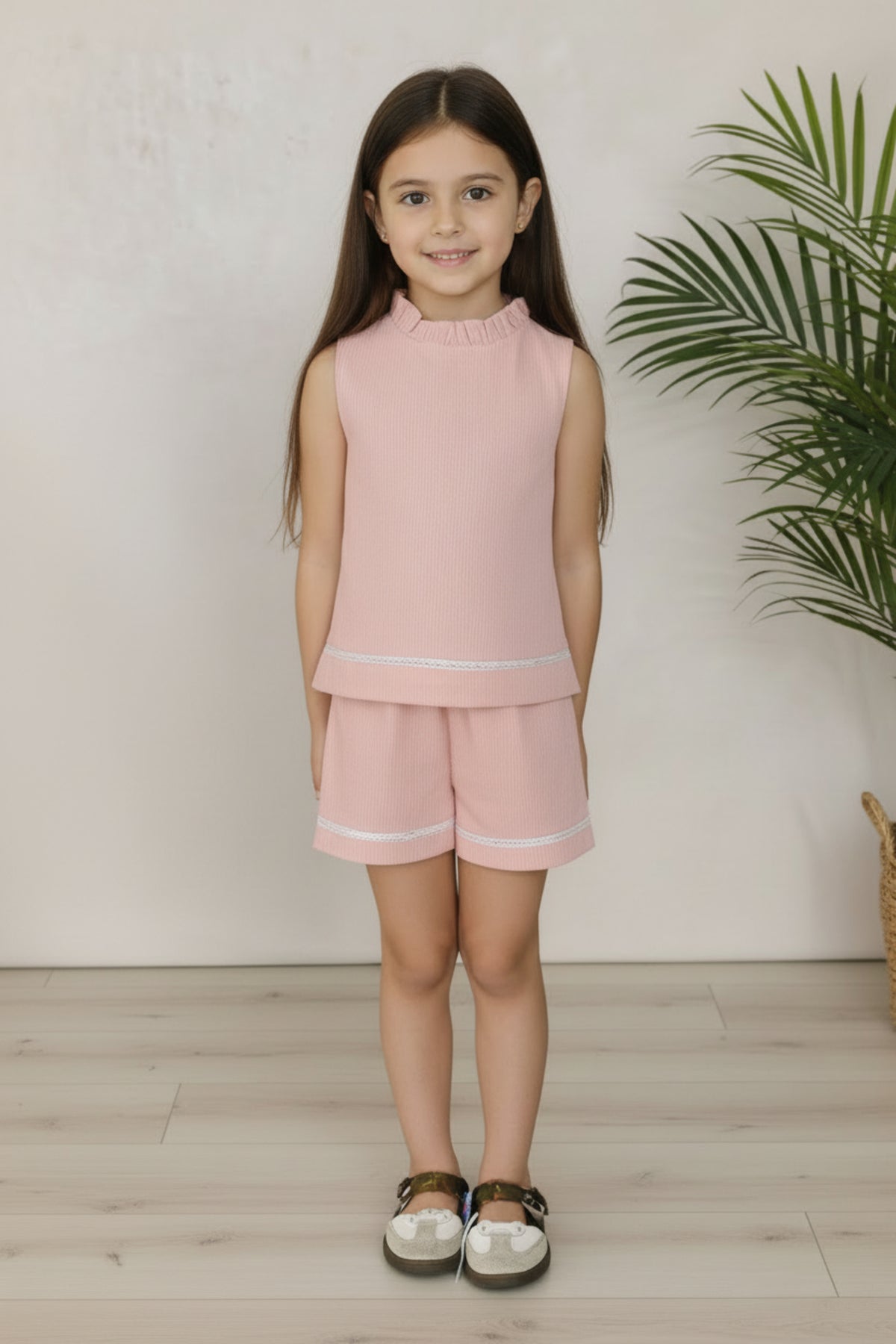 A young girl smiles while wearing a pink seersucker outfit with a ruffled neckline, paired with matching shorts and sandals.