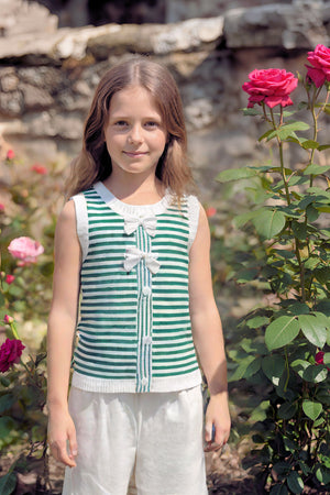 A young girl stands in a garden, wearing a green and white striped tank top with bows, complemented by light.