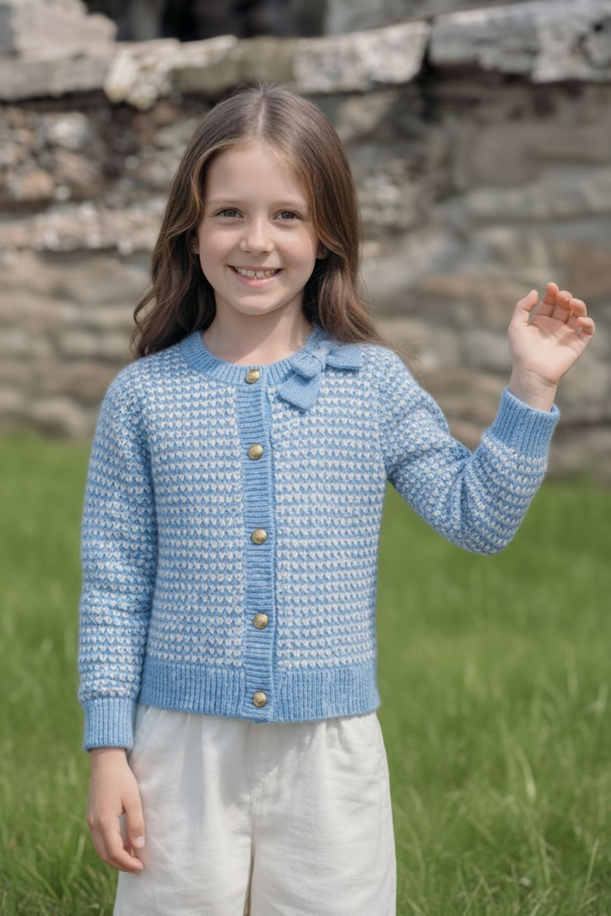 A smiling child wears a light blue cardigan featuring a textured tweed stitch and a bow detail at the collar, set against a grassy background.