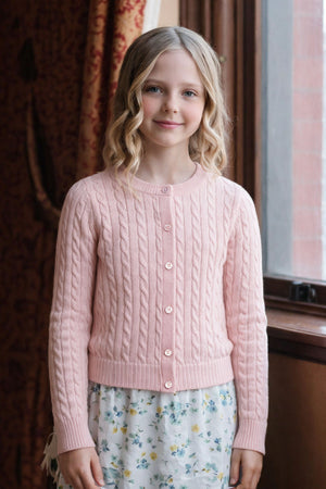 A girl wearing a soft pink cable knit cardigan with buttons stands near a window, showcasing a floral skirt.