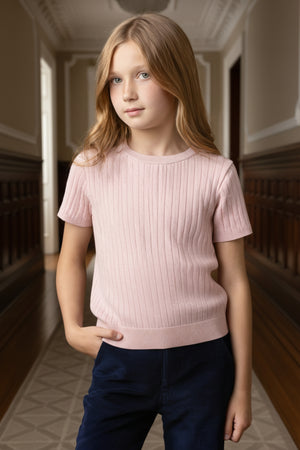 A girl stands confidently in a hallway wearing a light pink, short-sleeve cable knit sweater. The sweater, made from organic cotton, features a ribbed texture and a comfortable fit.