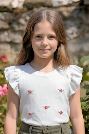 A girl is wearing a white ruffle sleeve sweater adorned with small floral embroidery, set against a lush garden background.