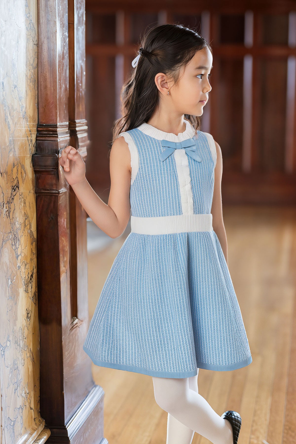 A young girl in a blue and white striped dress with a bow stands by a wooden column.