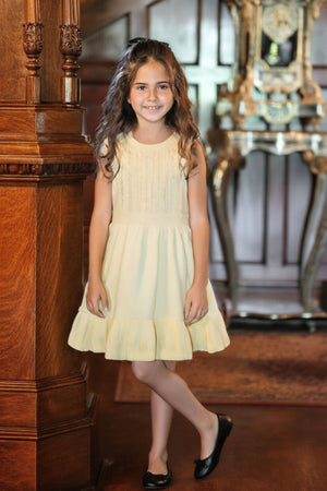 A young girl in a yellow flounce hem dress stands indoors, smiling, next to detailed wooden architecture.