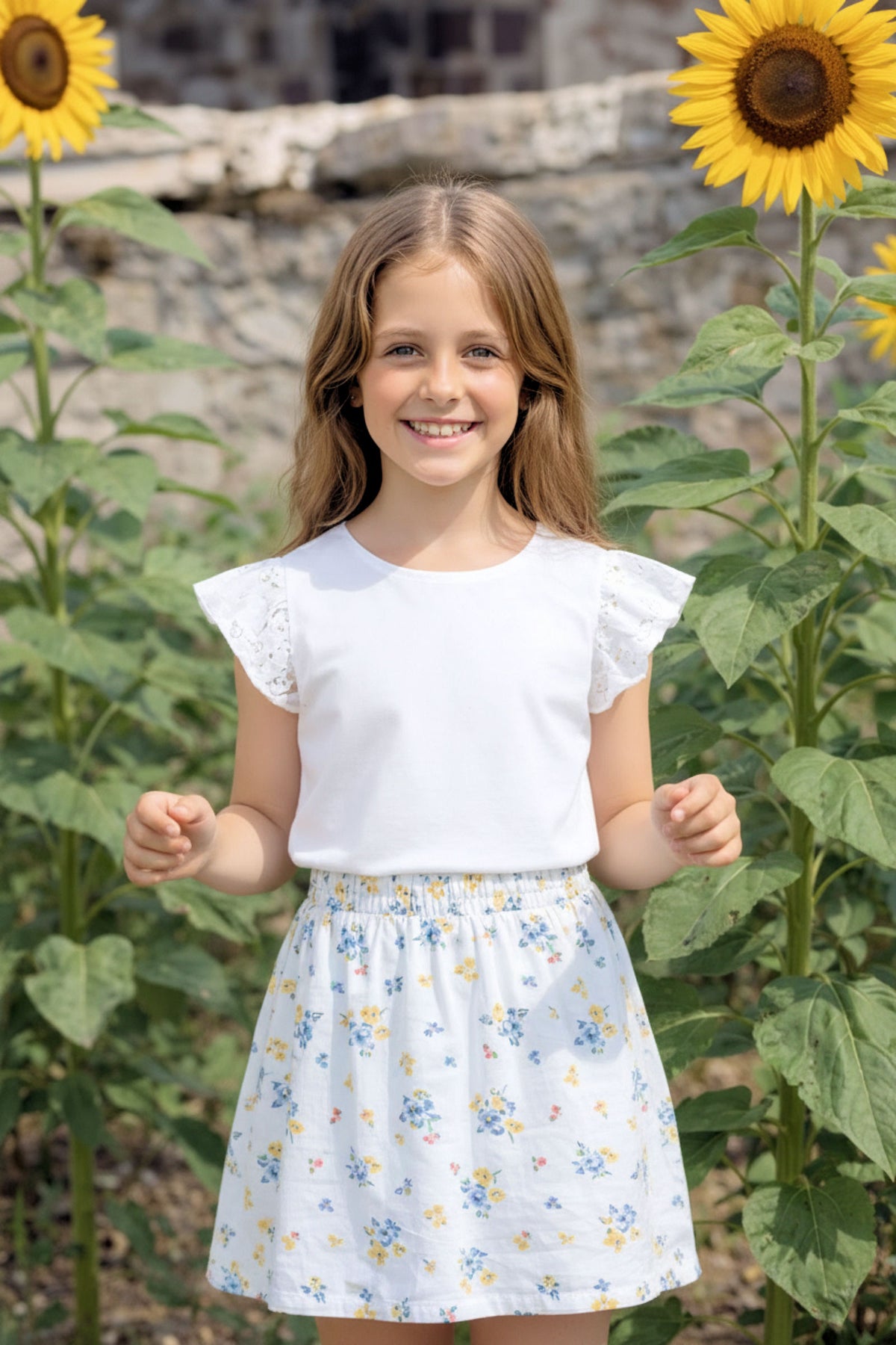 A girl smiles while wearing a white top with eyelet flutter sleeves and a floral skirt, standing among tall sunflowers.