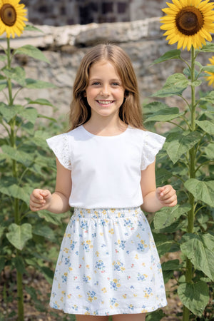 A girl smiles while wearing a white top with eyelet flutter sleeves and a floral skirt, standing among tall sunflowers.