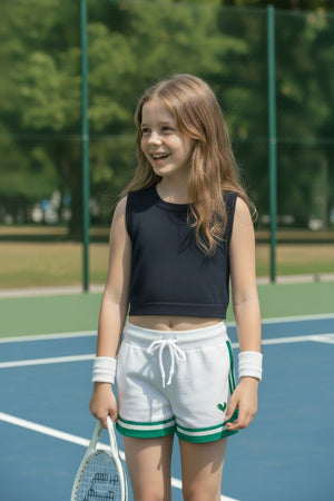A young girl stands on a tennis court wearing a navy crop top and white shorts with green athletic stripes.