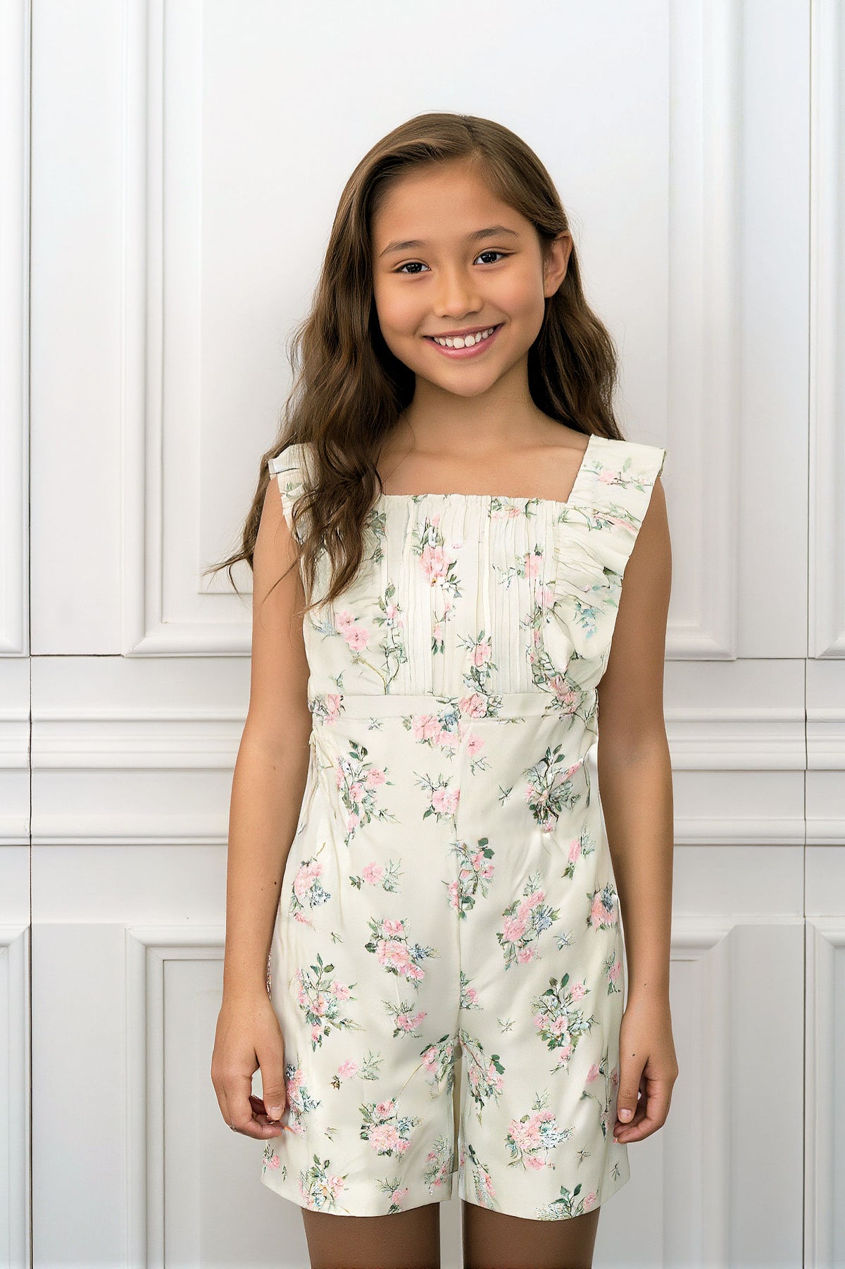 A girl smiles while wearing a floral-patterned jumpsuit with flutter sleeves, featuring a light cream background and pintuck details.