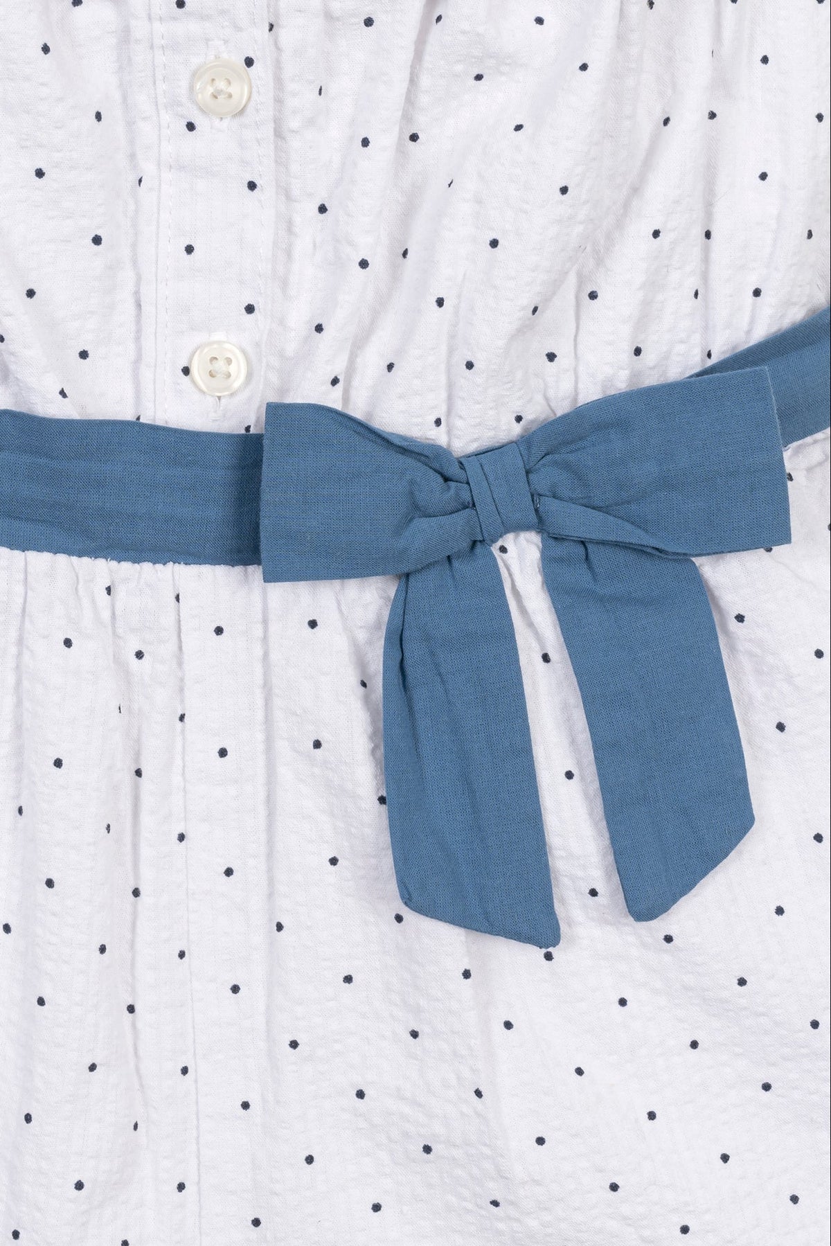 Close-up of a white seersucker romper featuring delicate blue dots and a matching bow tie at the waist. The fabric appears lightweight and perfect for warm weather wear.