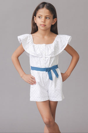 A young girl poses in a white organic cotton seersucker romper featuring a ruffle collar, blue waist sash, and side pockets, set against a neutral gray background.