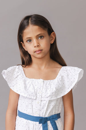 A young girl models a white organic cotton romper featuring a ruffle collar and a blue waist sash.