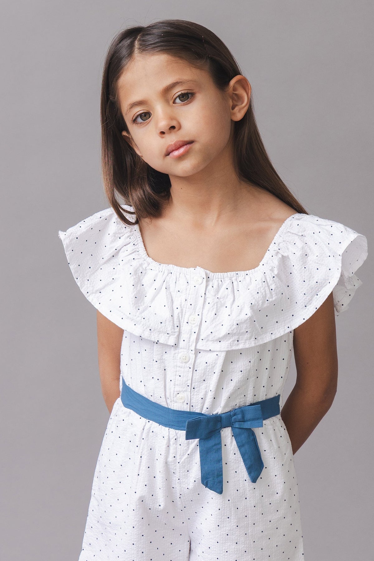 A young girl models a white organic cotton seersucker romper with a ruffle collar and a blue waist sash.