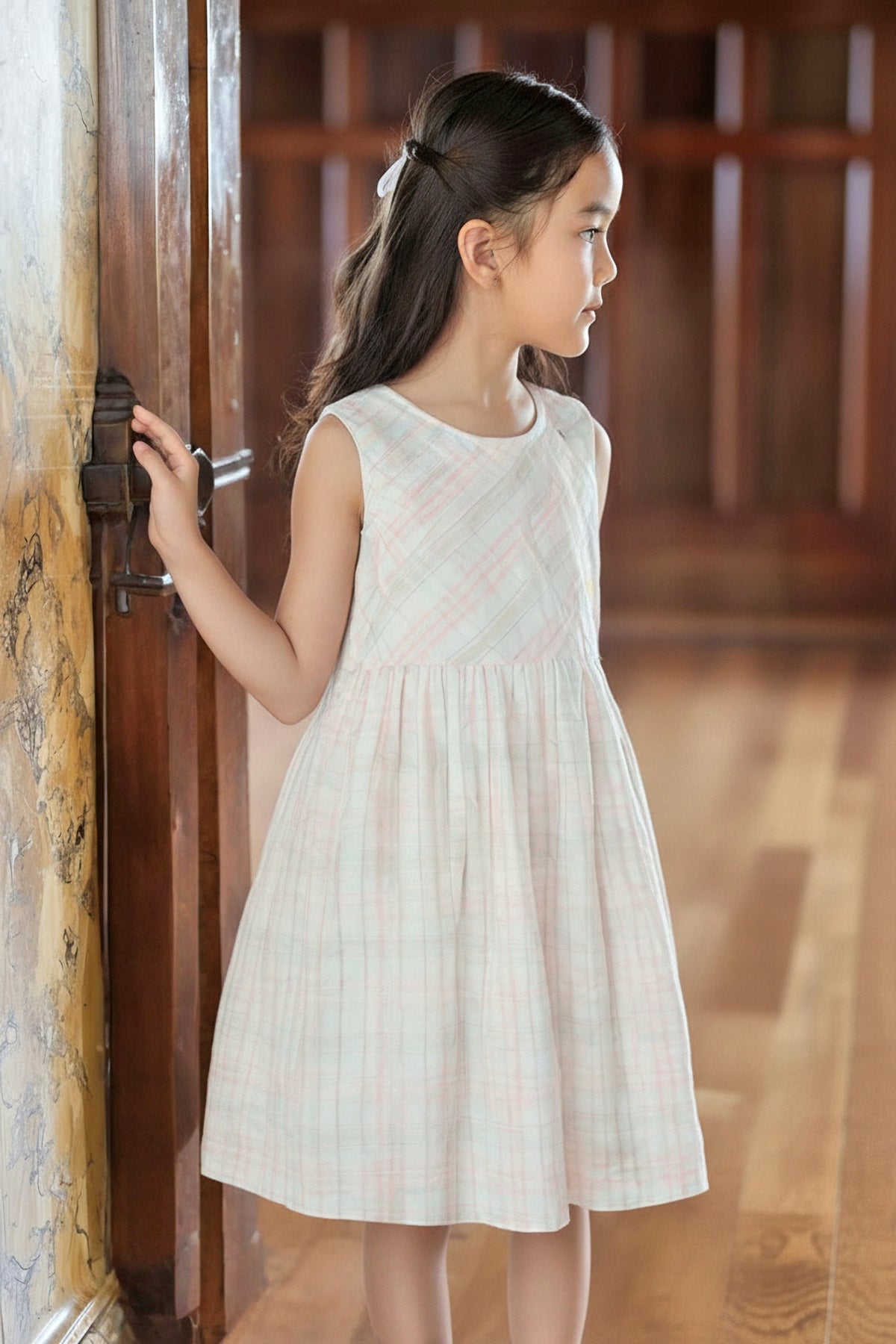 A girl wearing a sleeveless, plaid dress stands by a wooden door, showcasing soft pastel colors and a flared silhouette.