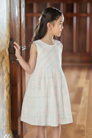 A girl wearing a sleeveless, plaid dress stands by a wooden door, showcasing soft pastel colors and a flared silhouette.