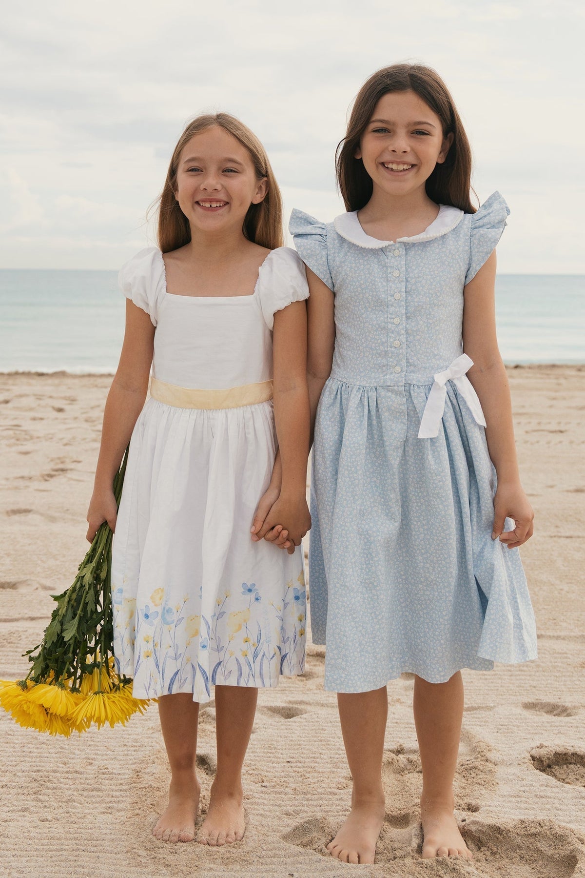 Two young girls stand barefoot on a sandy beach, holding hands. One wears a white dress with floral designs.