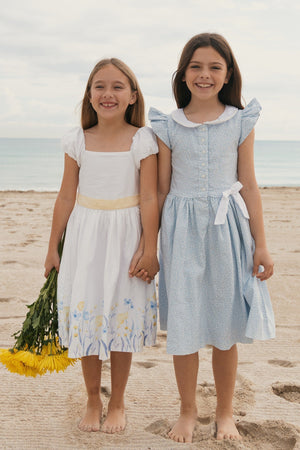 Two young girls stand barefoot on a sandy beach, holding hands. One wears a white dress with floral designs.