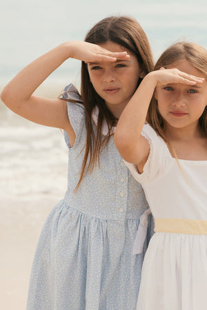 Two young girls stand on a beach, one wearing a blue linen dress with a Peter Pan collar and the other dressed in a white cotton dress, both smiling as.