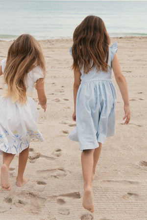 Two young girls walk barefoot on a sandy beach, one holding a bouquet of sunflowers. They wear charming dresses; one is white with floral details, and the other is light.